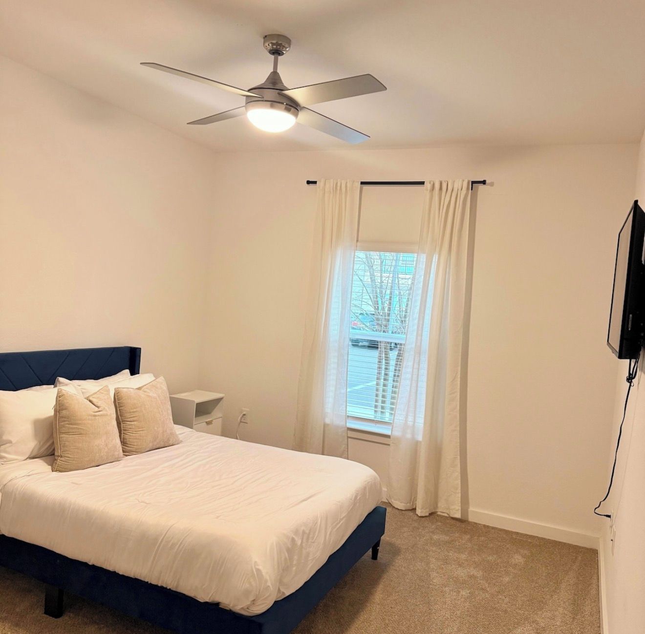 Un dormitorio con una cama, un ventilador de techo, una ventana con cortinas y un televisor montado en la pared.