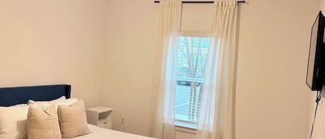 A bedroom with a bed, a ceiling fan, a window with curtains, and a television mounted on the wall.