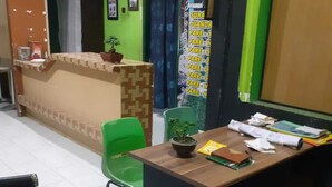 A reception desk with a green chair, a bulletin board with notices, and a wooden table with papers and a plant. - Nyaman Homestay (Pare)
