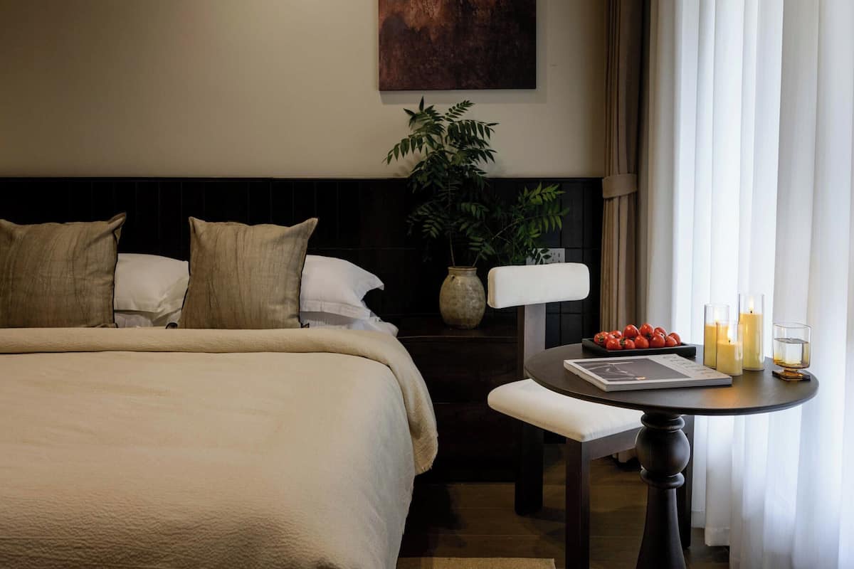 Ein Hotelzimmer mit einem großen Bett, einem Nachttisch mit Buch und Kerzen, einem kleinen Tisch mit Obst und einer Pflanze.