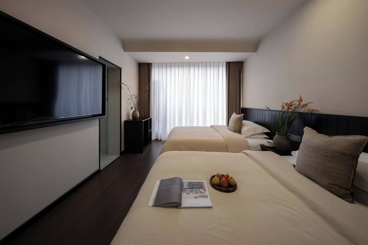 Ein modernes Hotelzimmer mit einem großen Flachbildfernseher, zwei Betten, einem Nachttisch mit einer Vase und einer Obstschale auf einem Bett.