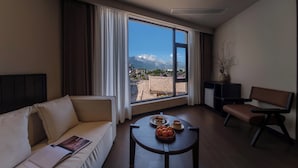 A modern hotel room with a large window offering a mountain view, a sofa, a small table with fruit, and a chair. - Lijiang Ancient City Hitone Resort (Lijiang)