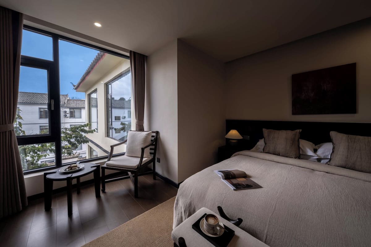 Ein Hotelzimmer mit einem großen Bett, einem Nachttisch mit Lampe, einem Sessel, einem kleinen Tisch mit Teller und Tasse sowie Blick auf Gebäude durch große Fenster.