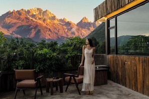 Une femme se tient dans une pièce d’où l’on peut admirer un paysage de montagnes. La pièce est dotée d’un mur en bois et d’une grande fenêtre. On y trouve des chaises et une petite table.
