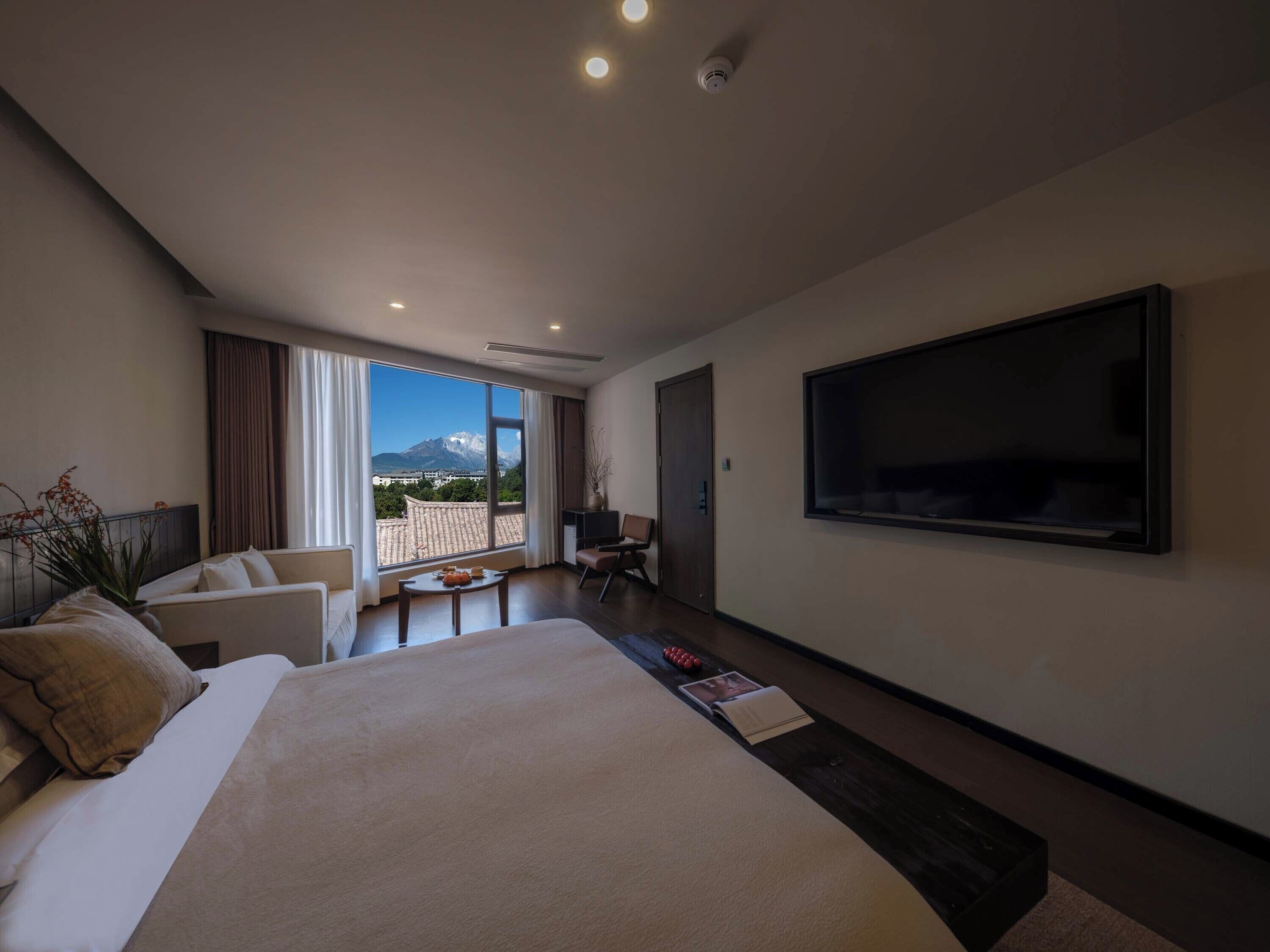 A modern hotel room with a large bed, a flat-screen TV, a dining area with a table and chairs, and a view of a mountain through large windows.