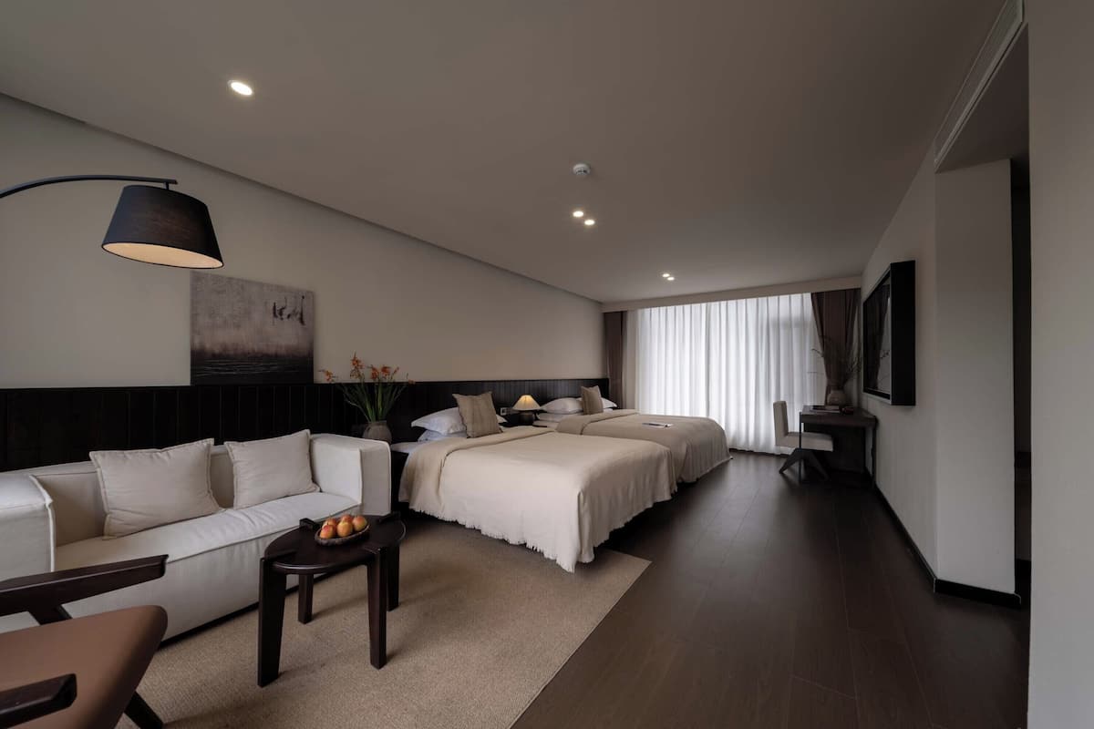 Ein modernes Hotelzimmer mit einer Couch, zwei Betten, einem kleinen Tisch mit Früchten, einer Lampe und einem Bild an der Wand.