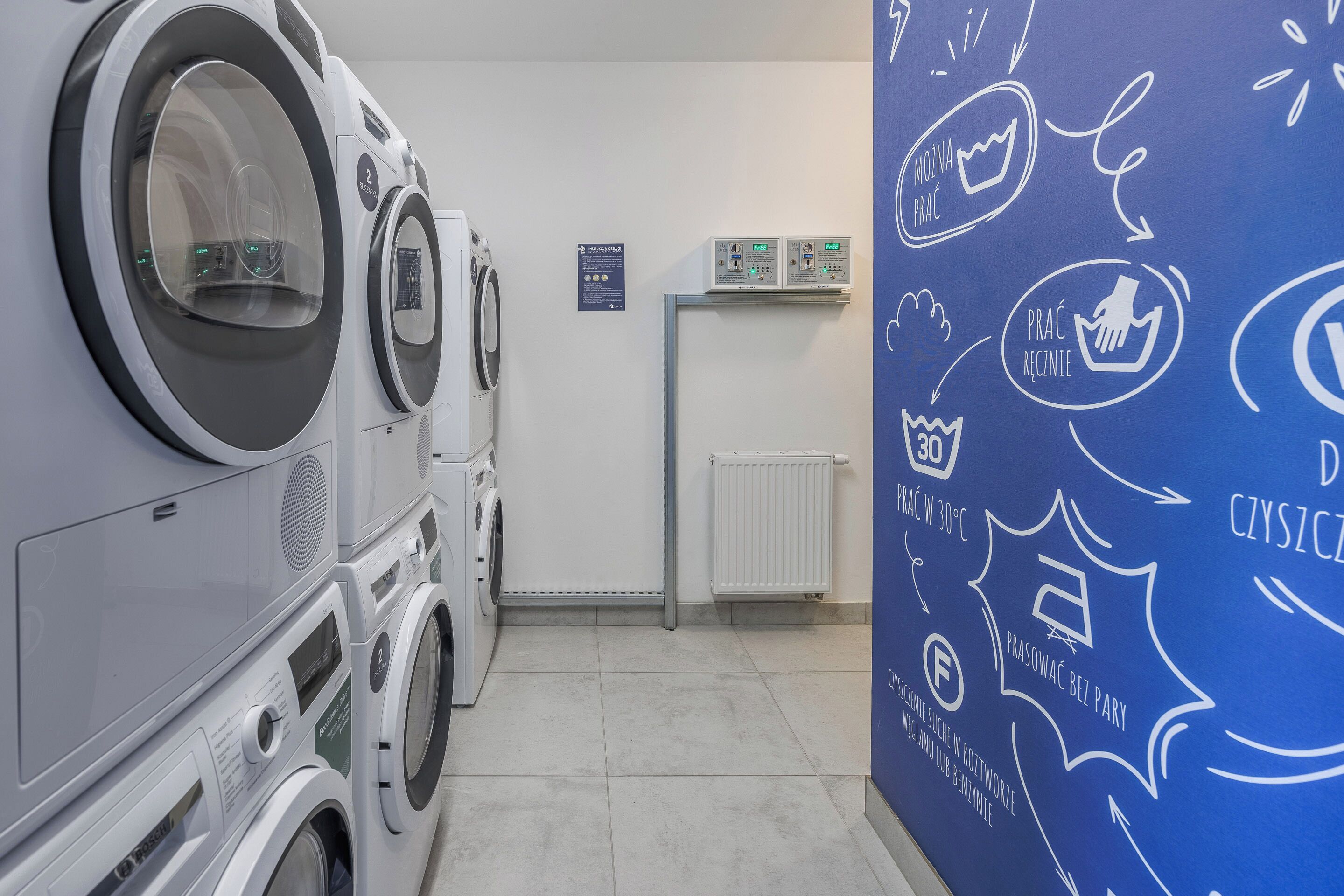 A laundry room with multiple washing machines and dryers, a white radiator, and a blue wall with laundry-related icons and text.