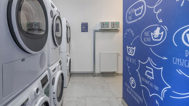 A laundry room with multiple washing machines and dryers, a white radiator, and a blue wall with laundry-related icons and text. - Emihouse Rubikon Apartments (Rzeszów)
