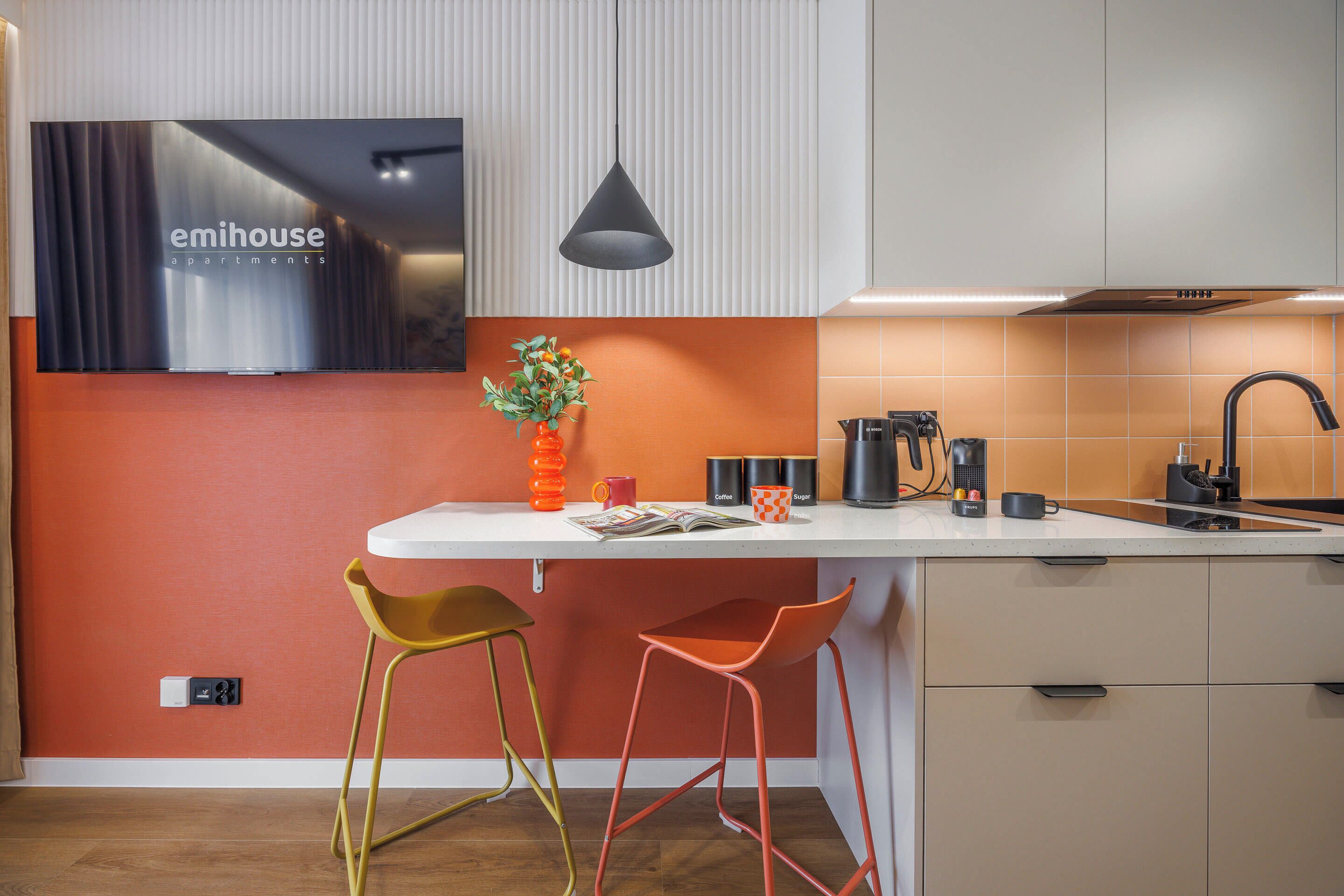 Una cocina moderna con encimera blanca, dos taburetes de bar, una lámpara colgante y el nombre de una marca visible en la pared.