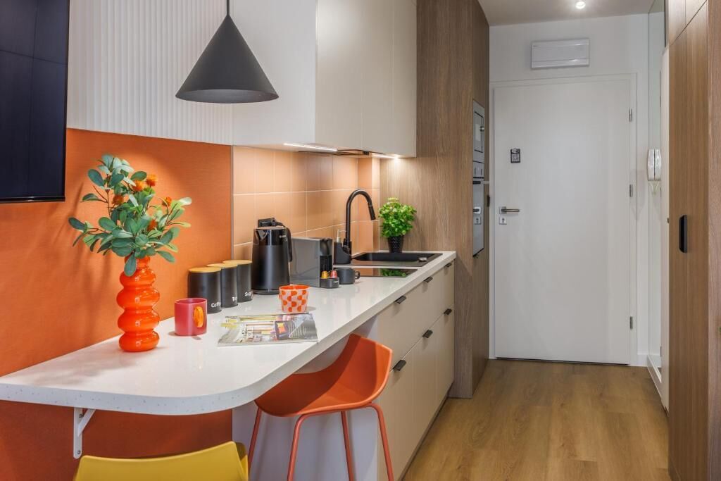 Una cocina compacta con encimera blanca, sillas anaranjadas, una lámpara colgante negra y una pequeña planta en maceta.