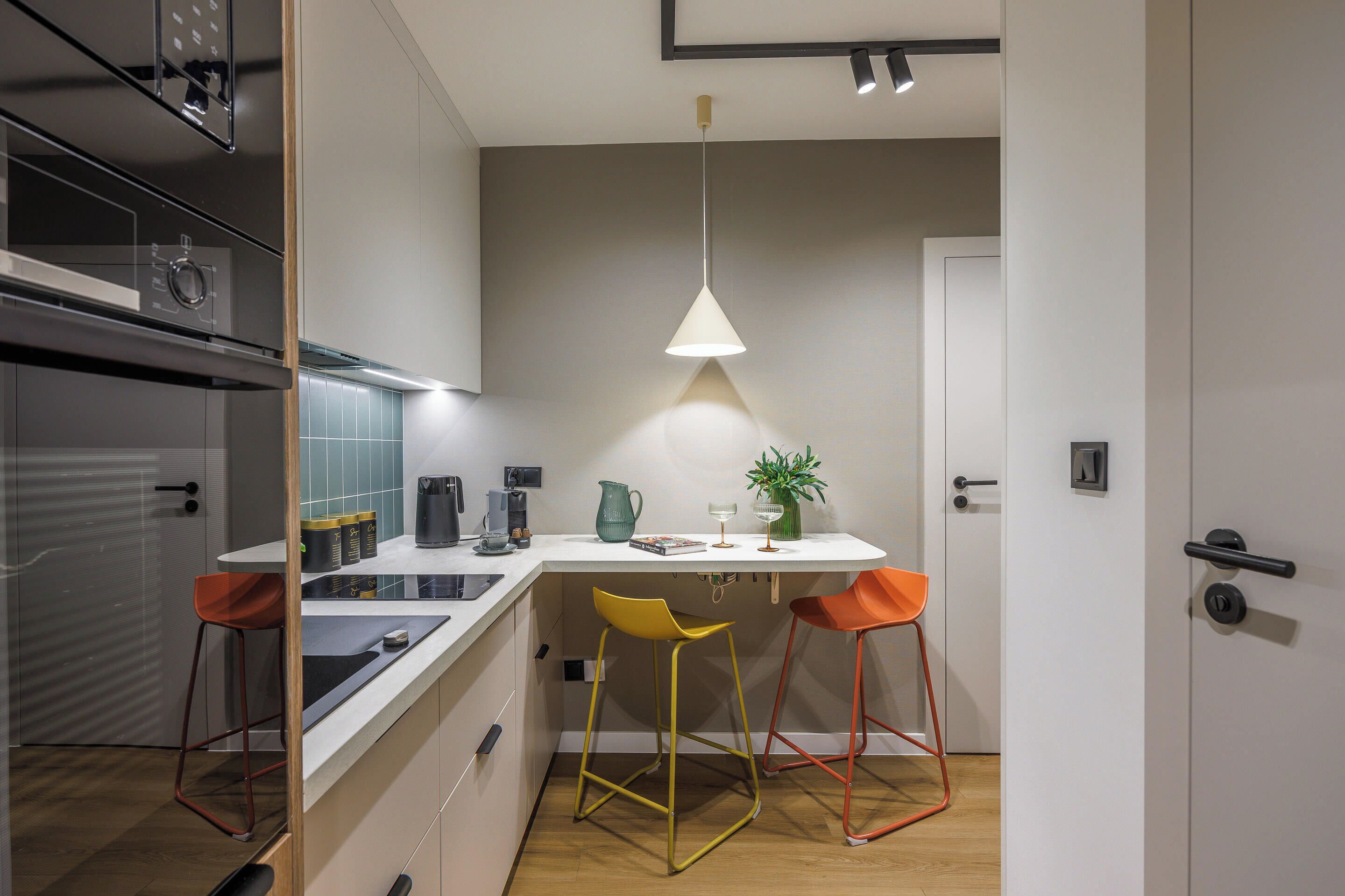 A compact kitchen with a white countertop, two stools, a hanging light, and a door.