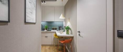 A narrow hallway leading to a kitchen with a white countertop, a sink, and a backsplash. There are two chairs with metal frames and orange seats.