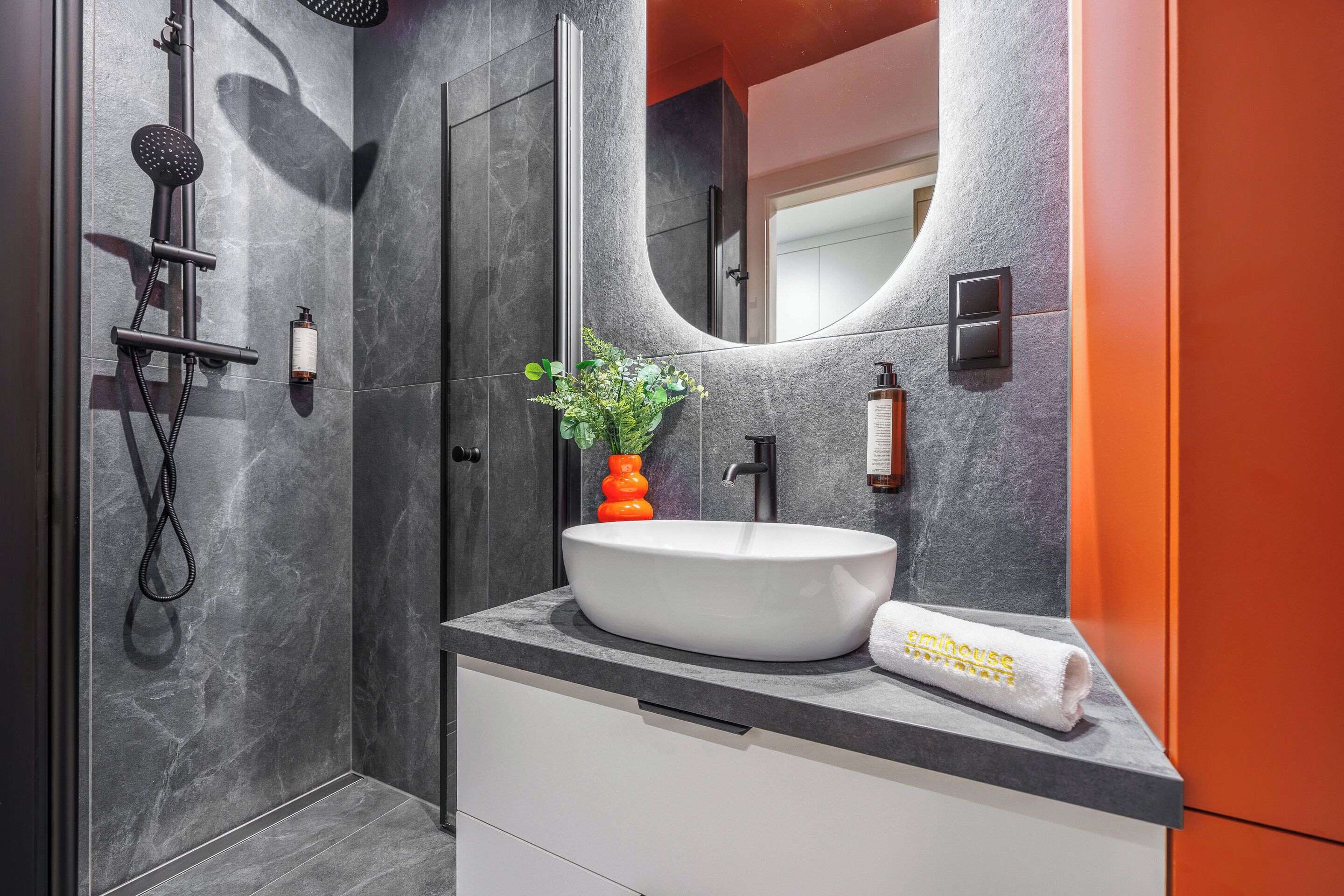A modern bathroom with a large mirror, a white sink, a showerhead, and a potted plant.