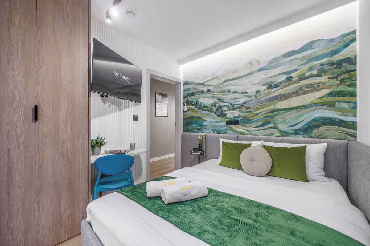 A hotel room with a bed, a desk, a chair, and a large landscape painting on the wall.