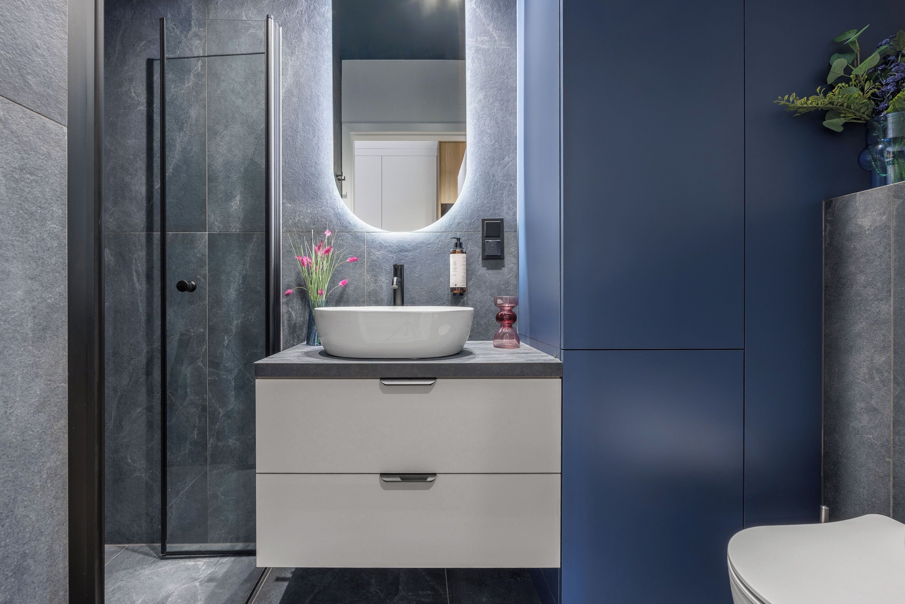 Ein modernes Badezimmer mit großem Spiegel, weißem Waschbecken, Duschkabine aus Glas und blauer Akzentwand.
