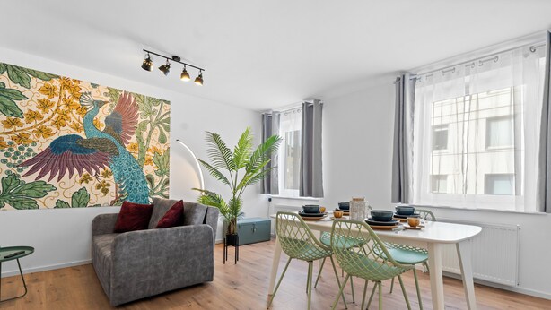A modern living room with a grey sofa, a dining table with chairs, a large window with curtains, and a colorful wall mural. - Central | Kitchen | Netflix | 43sqm (Remscheid)