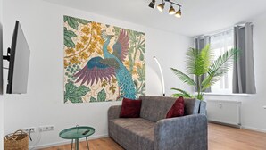 A modern living room with a grey sofa, a green glass table, a large floral artwork, a potted plant, and a window with curtains. - Central | Kitchen | Netflix | 43sqm (Remscheid)