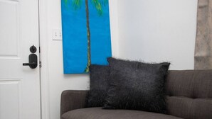 A room with a gray sofa, a black furry pillow, and a blue painting of palm trees on the wall. - Cozy Retreat | 5 mins to Beach & Downtown (Nassau)