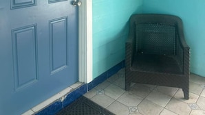 Eine blaue Tür mit kleinem Fenster und einem 'C' darüber, ein schwarzer Korbstuhl und eine wandmontierte Leuchte.