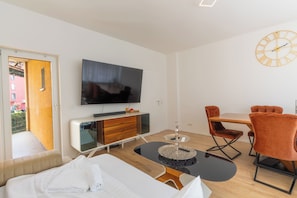 A modern hotel room with a bed, a TV, a dining table with chairs, and a clock on the wall.