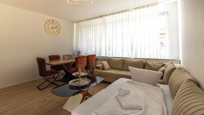 A modern hotel room with a wooden floor, a sofa, a dining table with chairs, a small round table with a glass top, a bed with white linens, a wall clock, and a large window with sheer curtains.