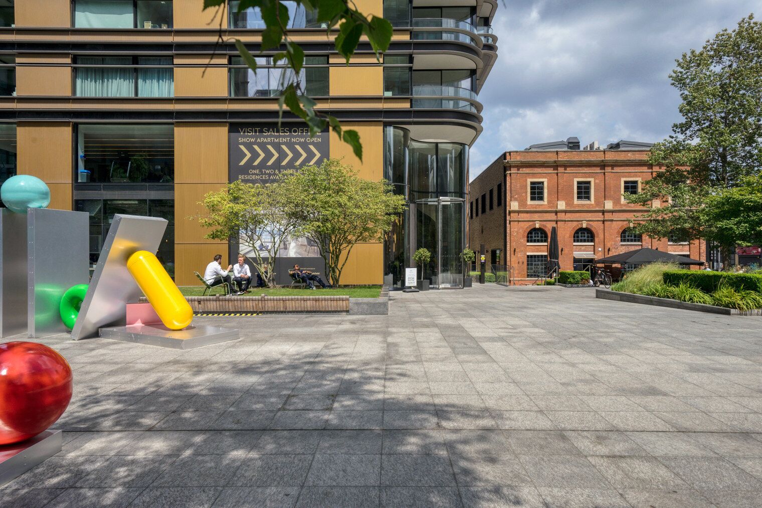 An open-plan office space with a modern building in the background, a glass elevator, and outdoor seating areas.