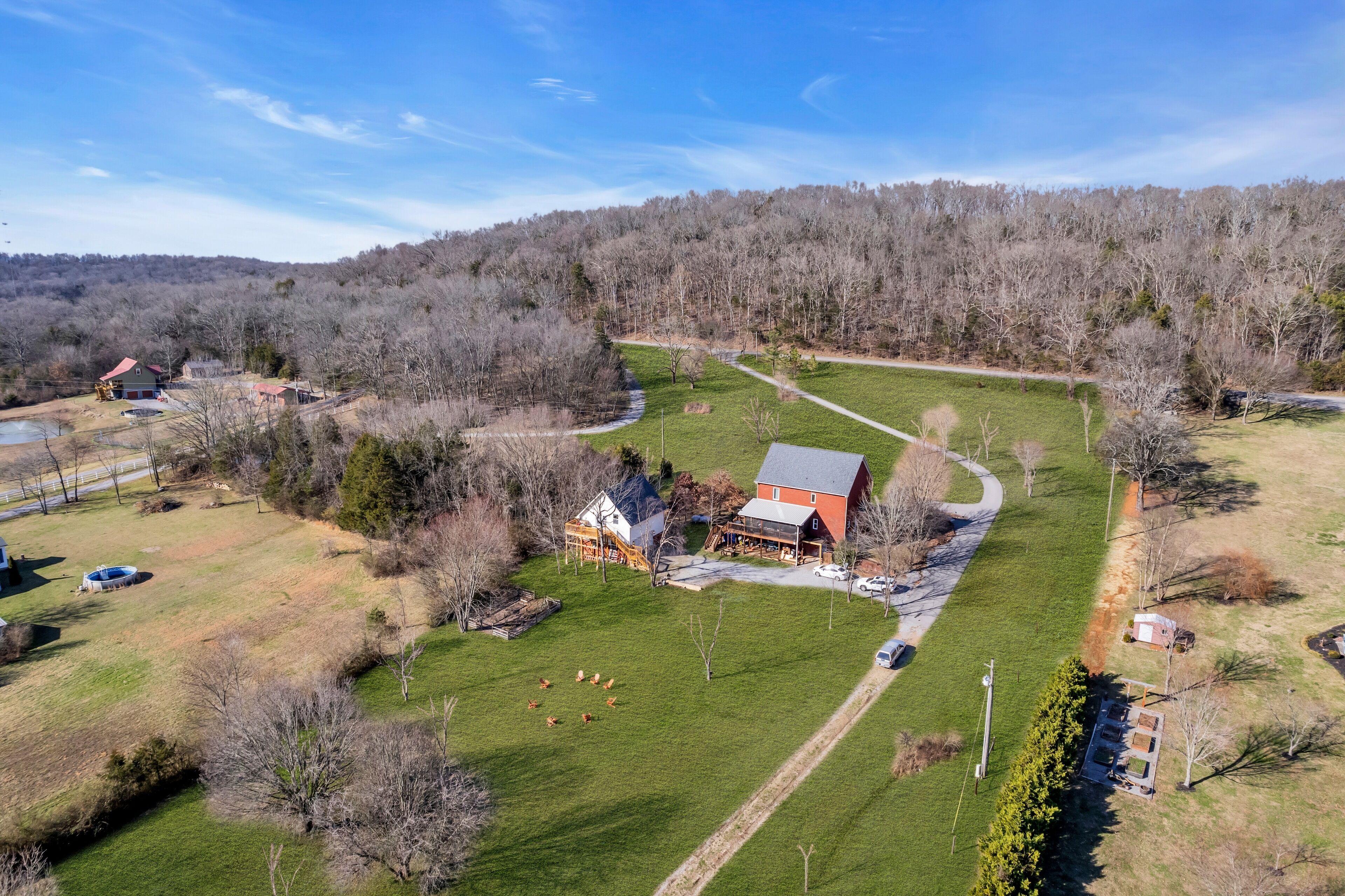 An aerial view of a rural property with a large house, a fenced yard, and a forested hill in the background.