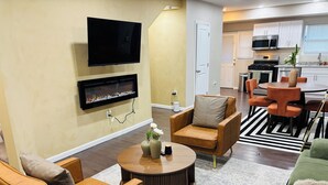 A living room with a flat-screen TV mounted on the wall, a fireplace below it, and a dining area with orange chairs and a round table. - Getaway in Greater Navy Yard - 4BR/3BA -Sleeps 10+ (Washington)