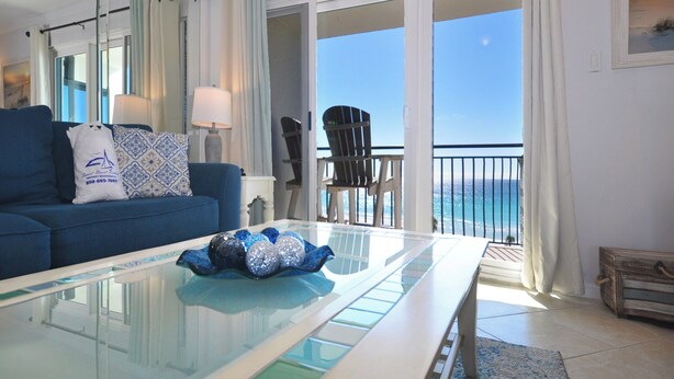 Ein Wohnzimmer mit Glastisch, blauem Sofa und einem Sessel. Große Glasfenster bieten Blick auf den Ozean.