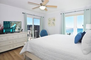Ein Schlafzimmer mit einem Bett, einem Fernseher auf einer Kommode, einem Deckenventilator und Meerblick durch große Fenster.