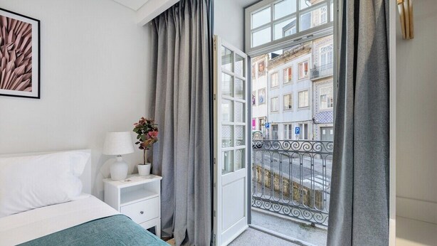 Ein Hotelzimmer mit einem Bett, einem Nachttisch, einer Lampe, einer Zimmerpflanze und Blick auf die Straße durch eine offene Tür.