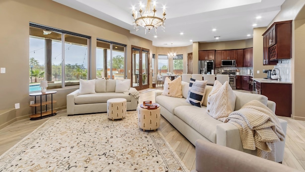 A spacious living room with a large sofa, ottomans, and a decorative rug. There is a kitchen area with modern appliances and wooden cabinets.