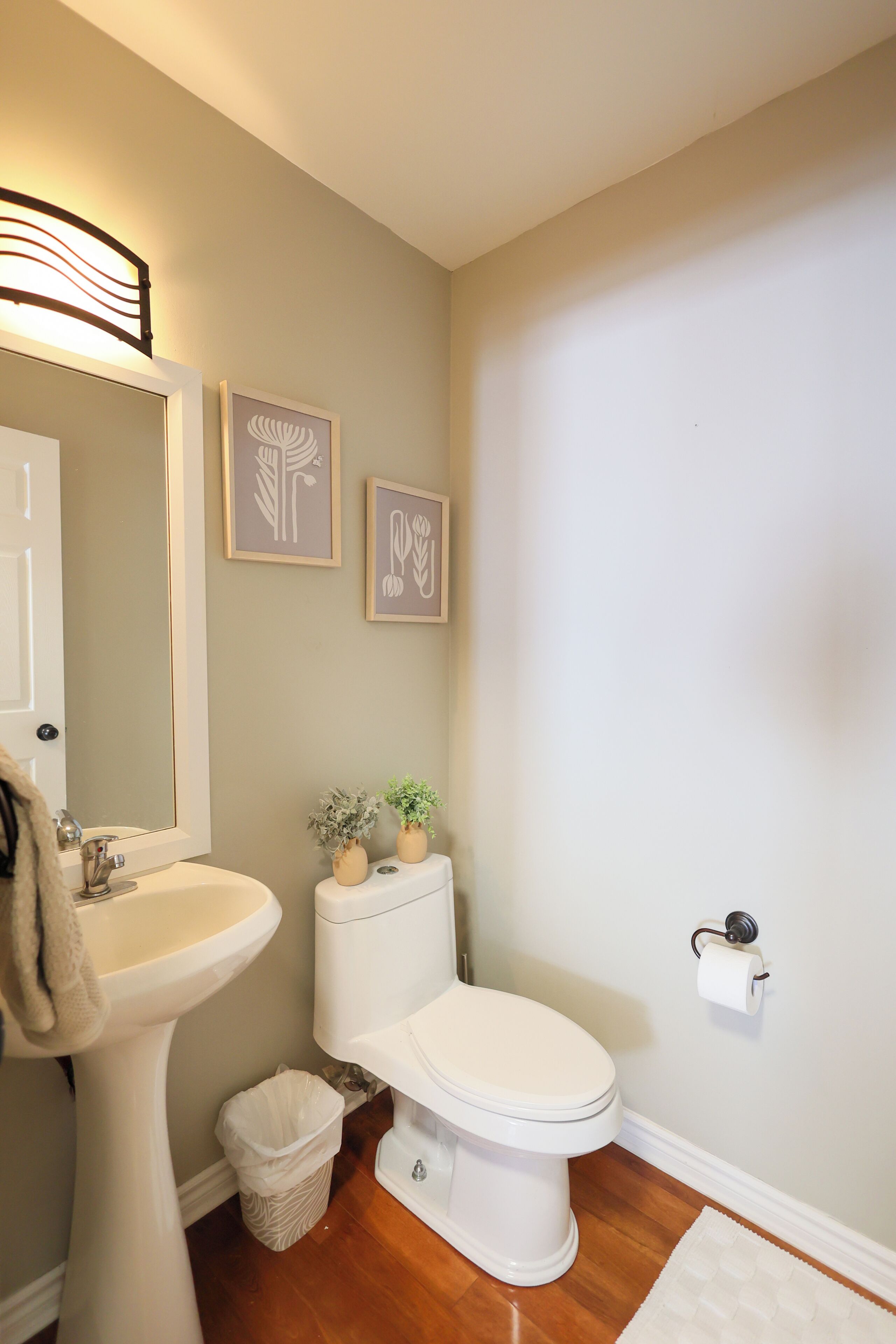 A bathroom with a white sink, toilet, and bidet, framed artwork on the wall, and a small trash bin.