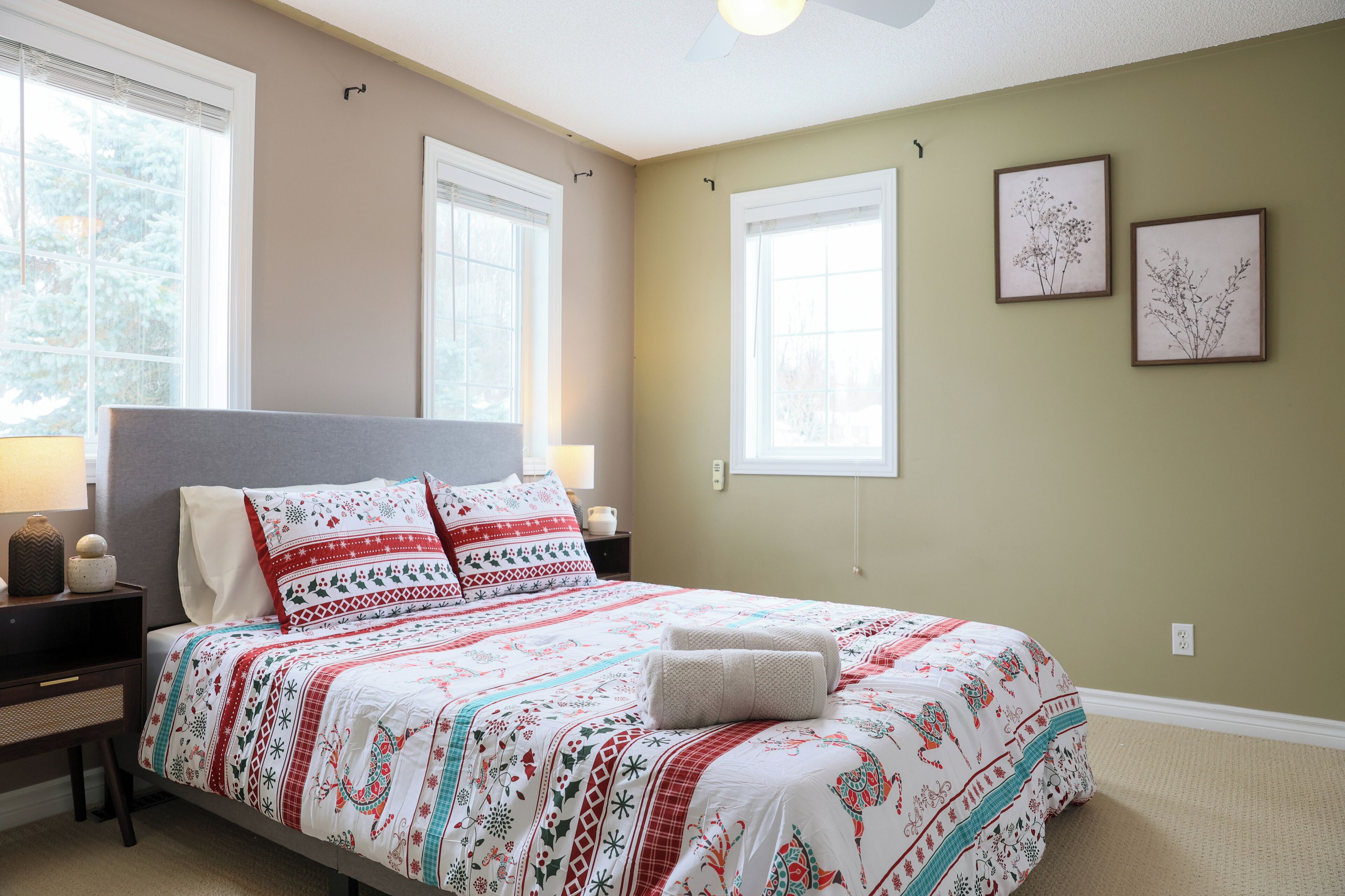 En pænt redt seng med et mønstret sengetæppe og puder, et natbord med en lampe, og to indrammede botaniske tryk på væggen.