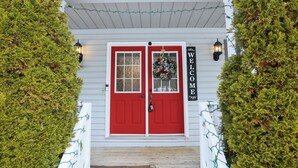 A red door with a wreath and a 'WELCOME' sign, flanked by evergreen trees and string lights.