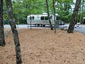 Un espacio recreativo con una caravana blanca estacionada entre árboles, un parque infantil y un sendero de grava.