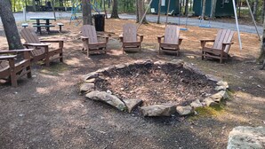 A campsite with a central fire pit surrounded by Adirondack chairs, picnic tables, and a playground in the background. - RV #1 - 30 Amp - Serene campground in Graysville perfect for outdoor adventures (Graysville)