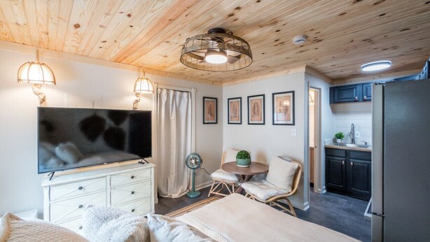 Ein gemütliches Zimmer mit einem Bett, einem Schreibtisch, einem Sessel, einem kleinen Tisch mit Pflanze, einem Fernseher und einer Küchenzeile.