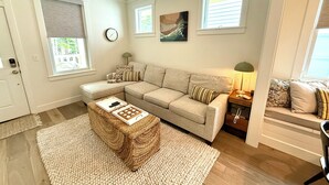 A living room with a beige sectional sofa, a coffee table with a board game, a clock on the wall, and a window with blinds.