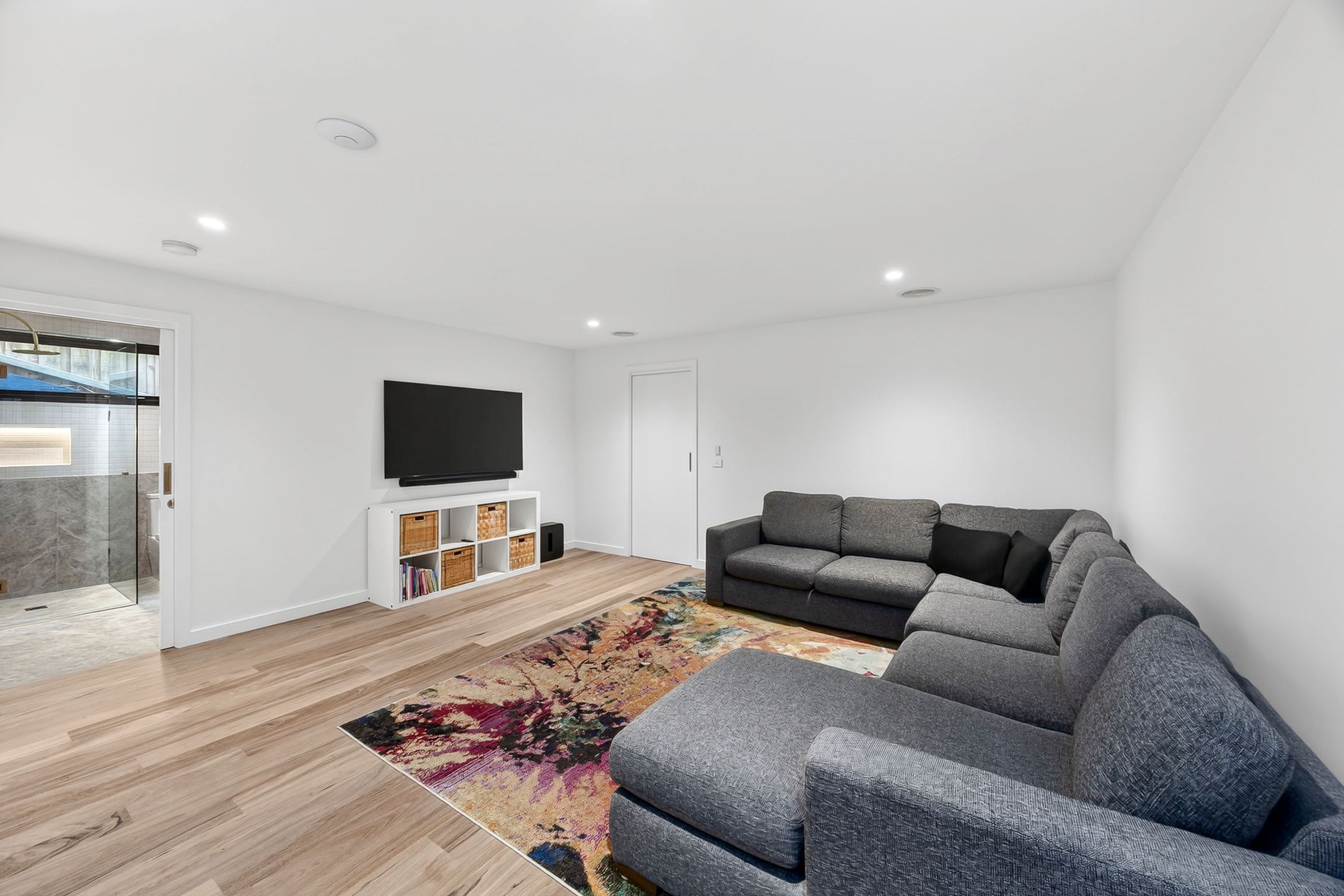 A modern living room with a grey sectional sofa, a flat-screen TV mounted on the wall, and a colorful rug on the wooden floor.