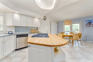 A modern kitchen with white cabinetry, a central island, and a dining area with wicker chairs. - Indigo Sky Oasis: Pool, Hot Tub, Near Joshua Tree (Yucca Valley)