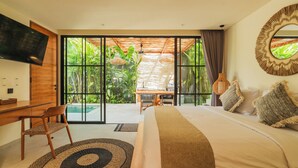 A bedroom with a bed, a desk, a chair, a TV, and a large window with a view of greenery. - Villa Rasa Rasa by Alfred in Bali (Cemagi)