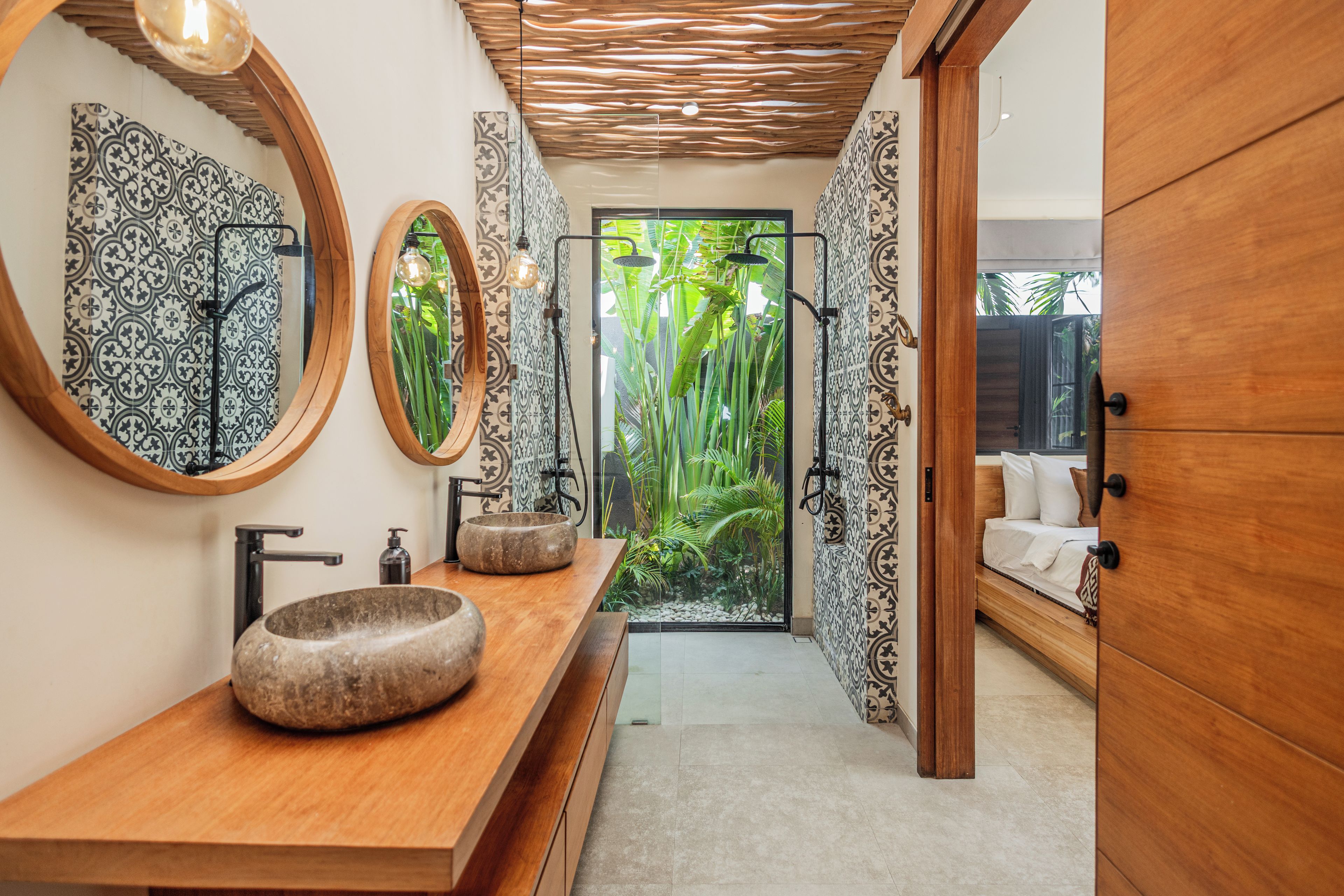 Een moderne badkamer met een houten wastafelmeubel, twee grote spiegels, een stenen wastafel en een douchegedeelte met uitzicht op tropische planten.