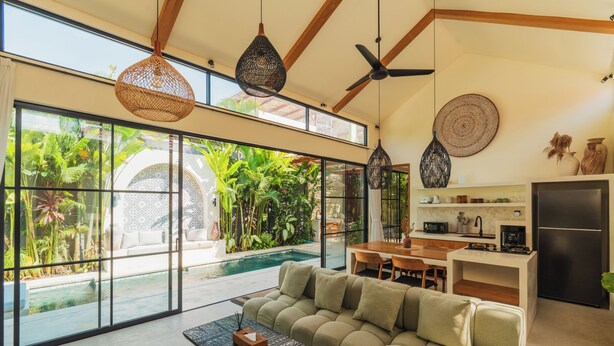A modern living room with a sofa, coffee table, and dining area. Large windows offer a view of a pool and lush greenery. - Villa Rasa Rasa by Alfred in Bali (Cemagi)