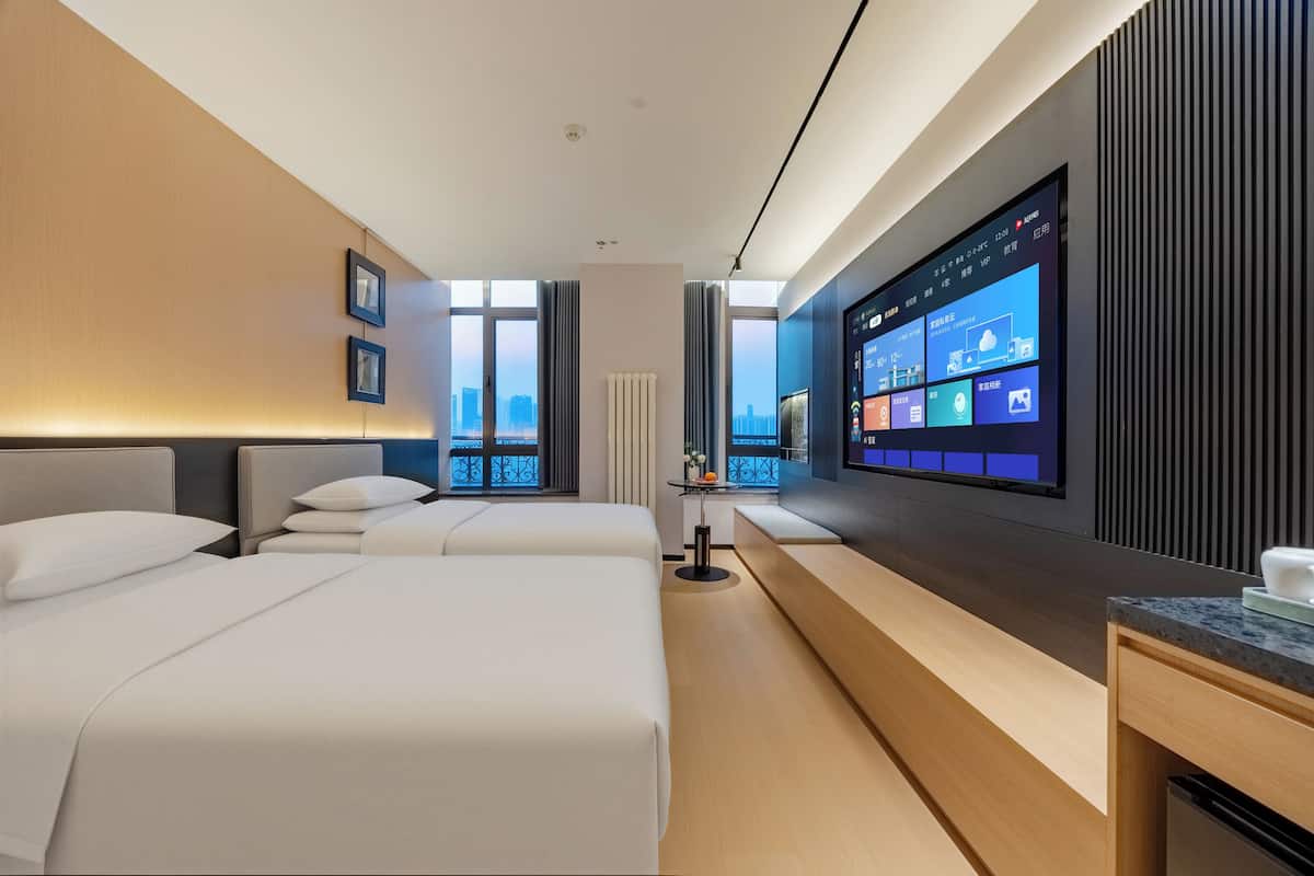 A modern hotel room with two beds, a large TV screen, and a view of the city.