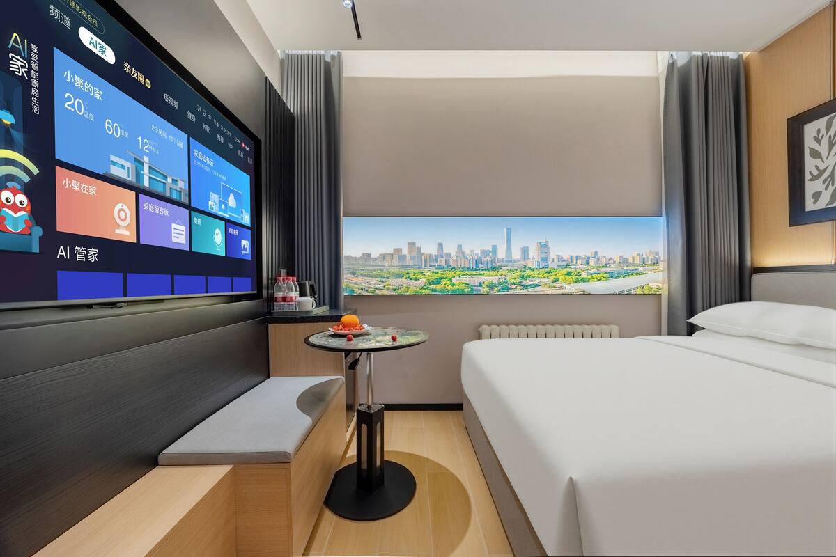 A modern hotel room with a large TV displaying a cityscape, a bed, a small table with fruit, and a view of the city through a window.