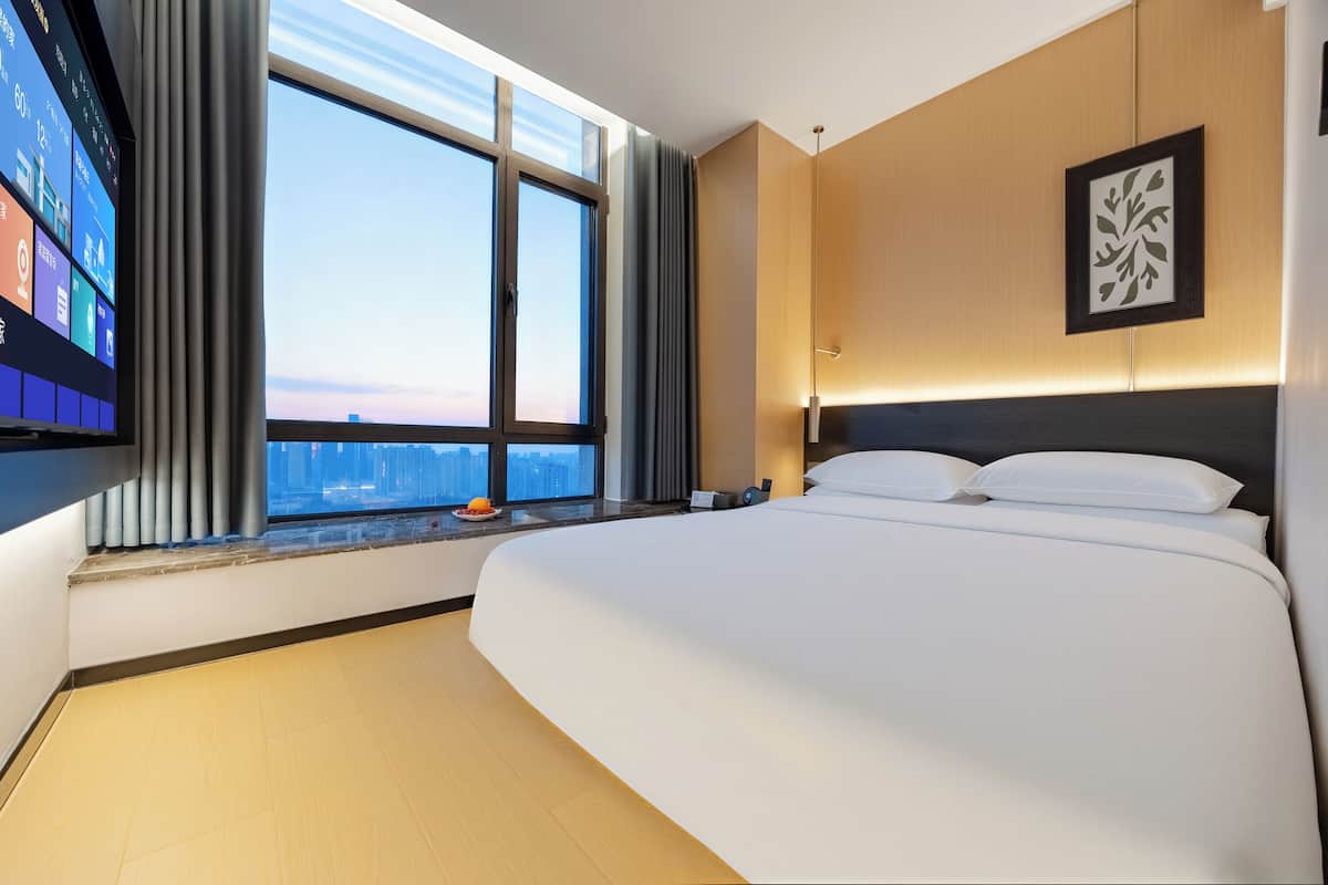 A hotel room with a large bed, a TV on the wall, a window with a city view, and a framed artwork on the wall.