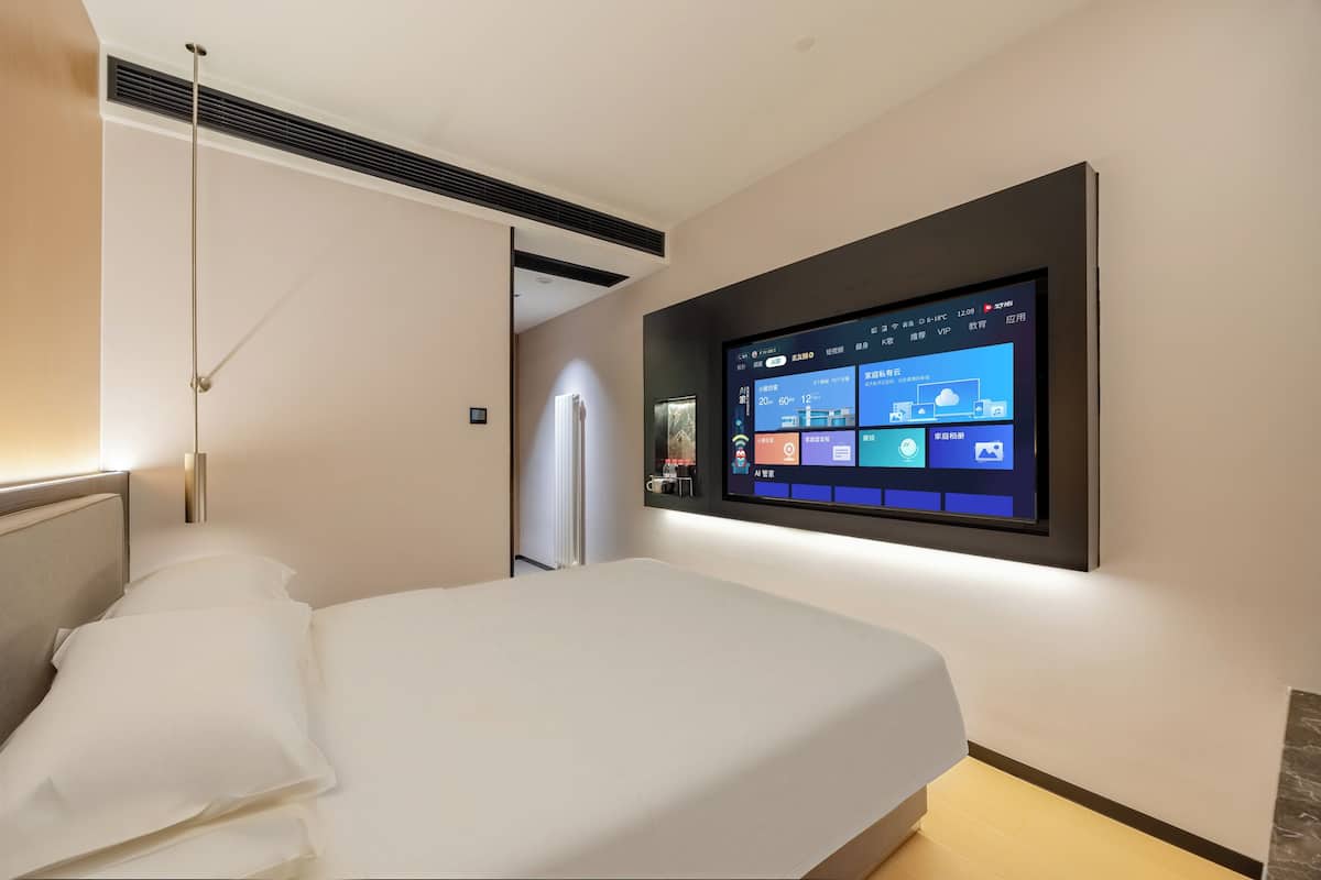 A hotel room with a bed, a mounted TV displaying a control panel, and a ceiling vent.