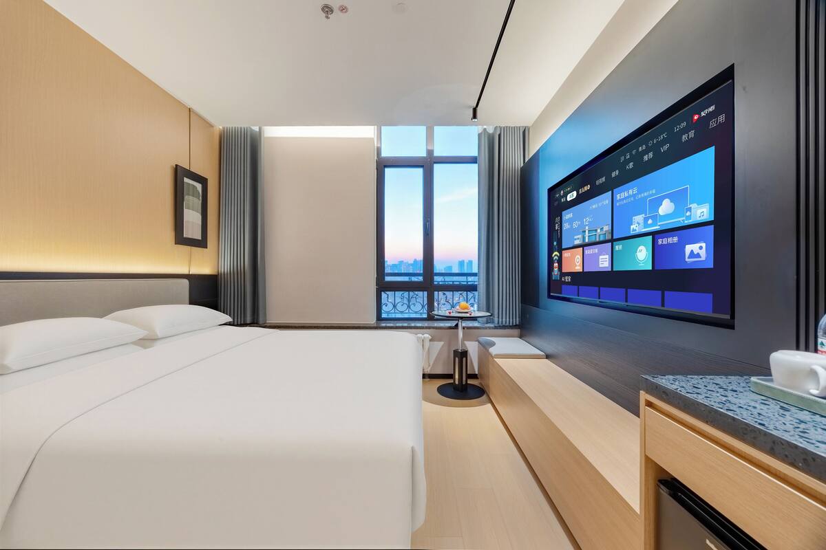 A modern hotel room with a large bed, a desk with a computer, and a view of the city through the window.