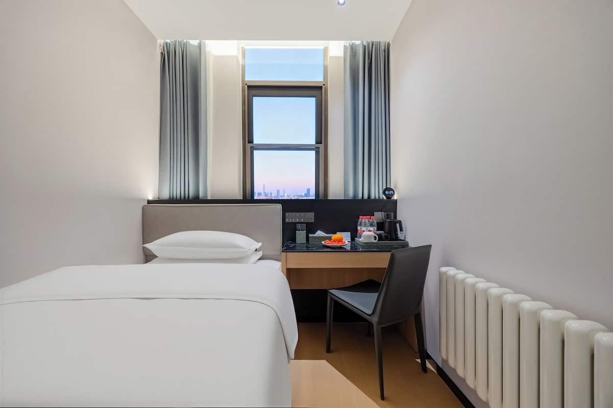 A hotel room with a bed, a desk, a chair, a window with curtains, and a radiator.